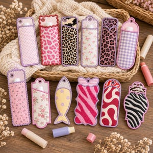 May include: A collection of twelve fabric lip balm holders featuring various patterns including pink polka dots, leopard print, floral motifs, purple gingham, zebra stripes, and abstract shapes, each with a small hole for attachment and stitched edges.