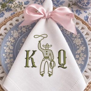 Puede incluir: Una servilleta de tela blanca con un bordado en color verde oliva que muestra a un vaquero lanzando un lazo entre las letras K y Q. La servilleta está atada en la parte superior con una cinta de satén rosa claro, formando un lazo decorativo.