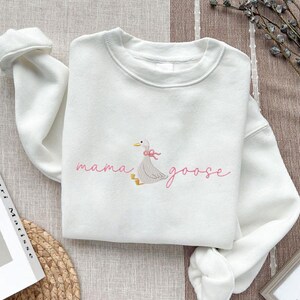 Peut inclure: Sweat-shirt blanc avec l'inscription brod&eacute;e "mama goose" en rose. Une oie grise avec un n&oelig;ud rose est brod&eacute;e &agrave; c&ocirc;t&eacute; du texte. Le sweat-shirt est en mati&egrave;re douce et poss&egrave;de un col rond.