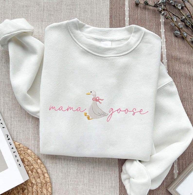 Peut inclure: Sweat-shirt blanc avec l'inscription brod&eacute;e "mama goose" en rose. Une oie grise avec un n&oelig;ud rose est brod&eacute;e &agrave; c&ocirc;t&eacute; du texte. Le sweat-shirt est en mati&egrave;re douce et poss&egrave;de un col rond.