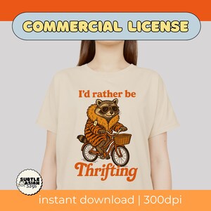 May include: Cream-colored t-shirt with a raccoon graphic wearing sunglasses and a tiger-striped coat, riding a bicycle with a basket. Text reads "I'd rather be Thrifting".