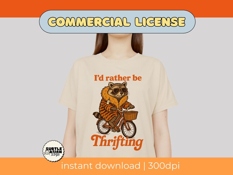May include: Cream-colored t-shirt with a raccoon graphic wearing sunglasses and a tiger-striped coat, riding a bicycle with a basket. Text reads "I'd rather be Thrifting".