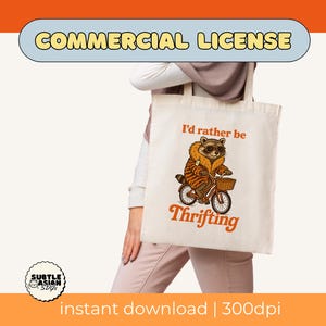May include: Cream-colored tote bag featuring a raccoon riding a bicycle with a basket. The raccoon wears sunglasses and a fur-trimmed coat. Text on the bag reads "I'd rather be Thrifting" in orange. The image includes the words "COMMERCIAL LICENSE".
