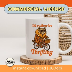 May include: White ceramic mug featuring a raccoon wearing sunglasses and a tiger-striped coat, riding a bicycle with a basket. The mug has the text "I'd rather be Thrifting" in orange. The mug sits on a wooden coaster.