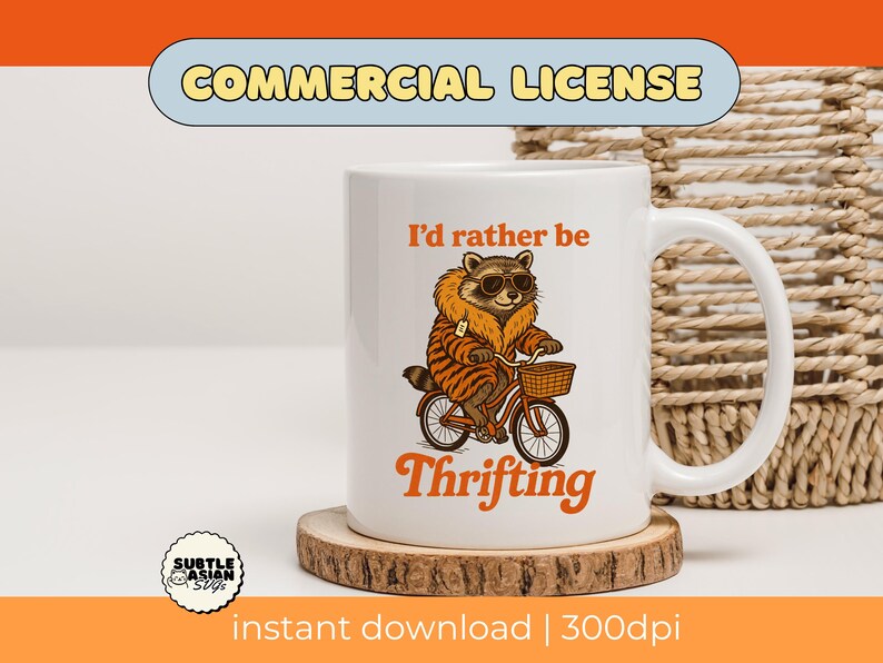 May include: White ceramic mug featuring a raccoon wearing sunglasses and a tiger-striped coat, riding a bicycle with a basket. The mug has the text "I'd rather be Thrifting" in orange. The mug sits on a wooden coaster.