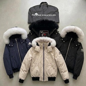 May include: Four Moose Knuckles bomber jackets with fur-trimmed hoods and a black garment bag. The jackets are navy, black, and beige, featuring front zips, metal hardware, and the Moose Knuckles logo on the sleeves. The garment bag is black.