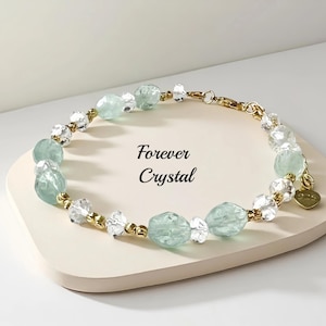 May include: A gold-toned bracelet with alternating clear and light green crystal beads. The bracelet is displayed on a white surface with the text "Forever Crystal". A small gold charm hangs from the bracelet.