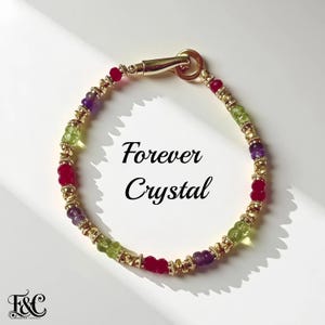 May include: A gold-toned bracelet with a variety of colorful gemstones, including red, green, and purple beads. The bracelet is adorned with gold accents and features the text "Forever Crystal".