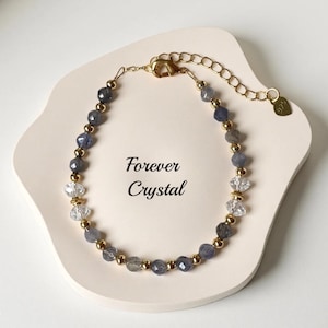 May include: A beaded bracelet with blue-gray and clear crystal beads, interspersed with gold-colored beads. The bracelet is on a cream-colored surface, with the text "Forever Crystal" in the center. The bracelet has a gold clasp.