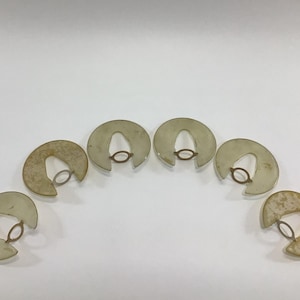 May include: Six semi-circular, off-white objects with a small, circular, bronze-colored ring in the center. The objects are arranged in a semi-circle against a white background. The objects have a slightly aged appearance.