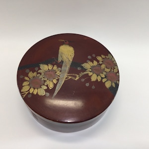 May include: A round, wooden box with a red lacquered finish. The lid is decorated with a gold and black painted design of a bird perched on a branch with flowers.
