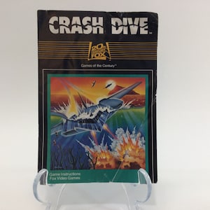 May include: A vintage game instruction booklet for Crash Dive, a video game from 20th Century Fox. The cover features a blue and gray fighter jet flying over a blue ocean with a volcano in the background. The text "Crash Dive" is in white letters on a black background.