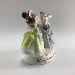 May include: A porcelain figurine of a mouse and a rabbit in a wedding scene. The mouse is wearing a light green dress with a yellow bow and the rabbit is wearing a light blue dress with pink flowers. The base of the figurine reads "The day I made you mine."