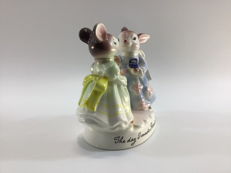 May include: A porcelain figurine of a mouse and a rabbit in a wedding scene. The mouse is wearing a light green dress with a yellow bow and the rabbit is wearing a light blue dress with pink flowers. The base of the figurine reads "The day I made you mine."