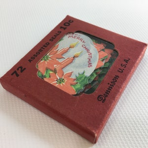 K&ouml;nnte beinhalten: Eine rote Schachtel mit 72 verschiedenen Weihnachtsaufklebern, die eine Weihnachtsstern und Kerzen mit dem Text "Merry Christmas" zeigen. Die Schachtel ist mit "72 Assorted Seals 10c Dennison U.S.A." beschriftet.