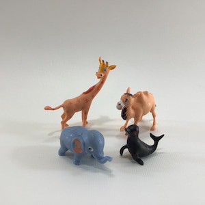 Könnte beinhalten: Ein Set aus vier Tierfiguren aus Plastik: eine Giraffe mit orangefarbenen und roten Markierungen, ein Kamel mit braunem Fell, ein blauer Elefant und eine schwarze Robbe.