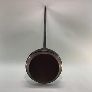 May include: A vintage copper pan with a long, curved handle. The pan is round and has a dark brown interior.