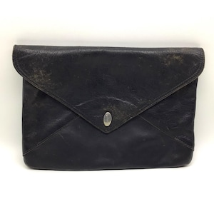 May include: A black leather envelope-style clutch bag featuring a pointed flap closure secured with a single oval-shaped silver-toned metal stud. The exterior surface displays a natural grain texture with visible signs of wear and light scuffing.