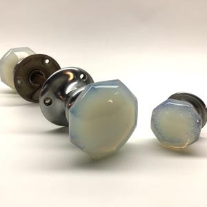 May include: Three octagonal door knobs featuring translucent, milky white glass faces with a smooth, faceted finish. Each knob is attached to a circular, metallic base plate with visible screw holes, showcasing a polished silver-toned metal hardware.
