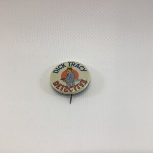 May include: A vintage Dick Tracy detective button with a red and white background. The button features a cartoon image of Dick Tracy holding a gun and wearing a blue suit and a yellow hat. The text "Dick Tracy Detective" is printed in blue around the image.