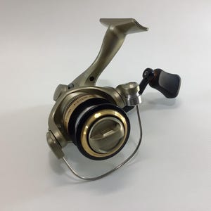 May include: A fishing reel with a metallic finish, featuring a handle, spool, and line guide. The reel has a black handle grip and gold accents around the spool. The design suggests it's for freshwater or saltwater fishing.