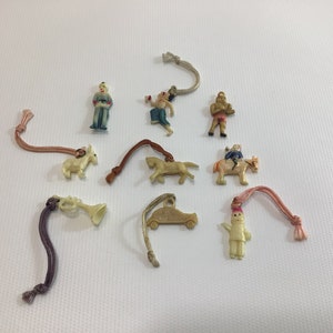 May include: A collection of ten vintage plastic charms with colorful details. The charms include a soldier, a man riding a horse, a car, a trumpet, a horse, a man with a cup, a man with a sword, a man with a flag, and a man with a hat. The charms are attached to strings.