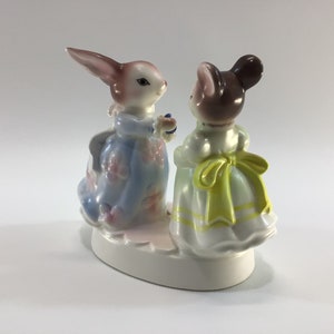 May include: A porcelain figurine of a rabbit and a mouse wearing dresses. The rabbit is wearing a blue dress with pink flowers and the mouse is wearing a white dress with a yellow sash.
