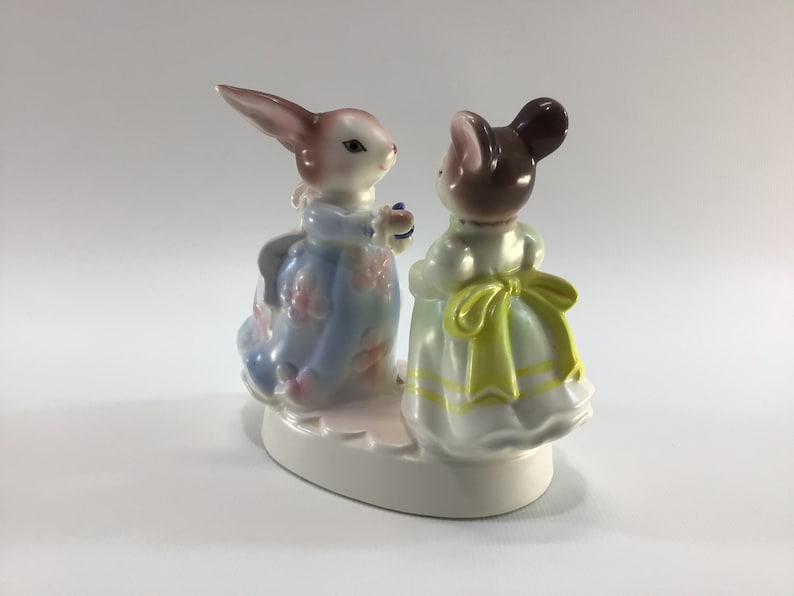 May include: A porcelain figurine of a rabbit and a mouse wearing dresses. The rabbit is wearing a blue dress with pink flowers and the mouse is wearing a white dress with a yellow sash.