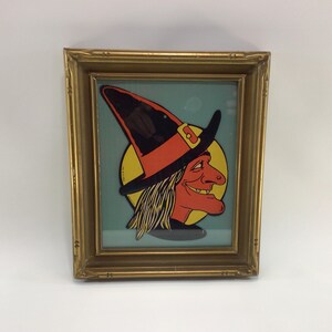May include: Framed vintage Halloween artwork featuring a cartoon witch's face. The witch has a red face, orange hat with a black brim, and yellow hair. The artwork is set against a light blue background and framed in a gold-colored frame.