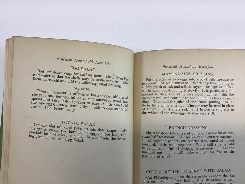 May include: A vintage cookbook with a yellowed, cream-colored paper cover. The book is open to two pages with the titles "Egg Salad", "Potato Salad", "Mayonnaise Dressing", "French Dressing", and "Cheese Relish to Serve with Salad". The text is in black ink and describes how to make each salad.