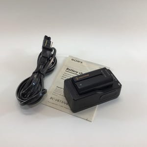 May include: A black Sony battery charger with a black power cord and a printed manual. The charger has a rectangular design with a battery slot on top. The manual is white with black text.