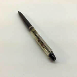 May include: A ballpoint pen featuring a glossy black tapered tip and a clear, transparent barrel that reveals internal mechanical components. The pen includes a silver-toned metal clip and a solid black cap at the end, creating a sleek design.