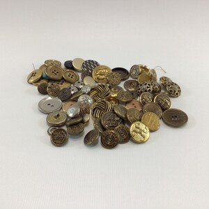 May include: A collection of assorted buttons in various sizes and designs. The buttons are primarily in shades of gold, bronze, and silver, with some featuring intricate patterns and textures. The buttons are scattered on a white surface.