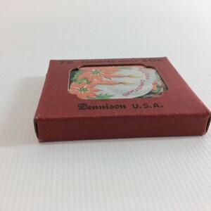 K&ouml;nnte beinhalten: Eine rote Schachtel mit einem durchsichtigen Fenster, das eine Bogen mit 72 verschiedenen Weihnachtsbriefmarken zeigt. Die Schachtel tr&auml;gt die Aufschrift "72 Assorted Seals 10c" und "Dennison U.S.A.". Die Briefmarken zeigen einen Weihnachtsstern und Kerzen mit dem Text "Merry Christmas" und "Merry Xmas".