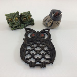 May include: Three owl figurines, two green and one gray, with a black metal owl trivet. The green owls are sitting side by side, while the gray owl is standing upright. The metal trivet is in the shape of an owl with a detailed design.