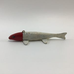 May include: A vintage fishing lure shaped like a fish. The lure has a red head and a white body with two small fins and a tail fin. A small metal loop is attached to the top of the lure.