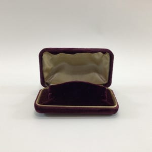 May include: An open, rectangular jewelry box with a deep burgundy velvet exterior and a tan satin interior. The box is designed to hold a ring or small piece of jewelry. The box is empty and sits on a white surface.