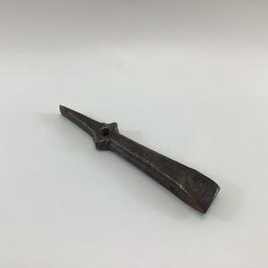 May include: A dark, weathered metal tool with a pointed end and a flat, beveled edge. The tool has a central hole and a textured surface, suggesting it is an antique or vintage item. The tool is likely used for prying or shaping materials.