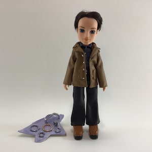 May include: A brown-haired doll wearing a brown jacket, a blue shirt, black pants, and brown shoes. The doll is standing on a white background. The doll is holding a purple tray with silver rings.