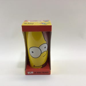May include: A yellow "Hi Ball Glass" featuring Homer Simpson's face, packaged in a red and yellow box. The glass has a cylindrical shape and is decorated with Homer's large eyes and smiling face.