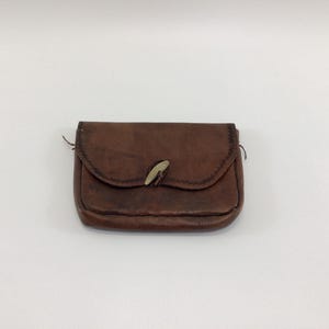 May include: A small, brown leather pouch with a curved flap and a light-colored clasp. The pouch has a stitched border and appears to be handmade. The leather has a worn, textured appearance, suggesting age or use. A simple, rustic accessory.