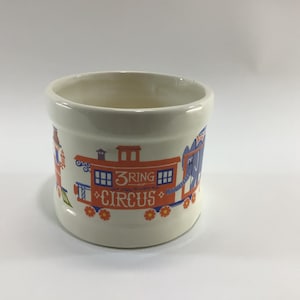 May include: Cream-colored ceramic mug with a circus train design. The train car is orange with blue and white accents, and the text "3 RING CIRCUS" is displayed on the side. The mug has a wide, open top and a simple, rounded shape.