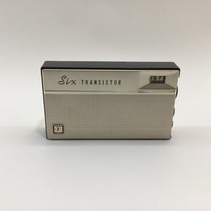 May include: A vintage transistor radio with a silver and black design. The radio has the words "Six Transistor" in red script and a small display. The front of the radio has a speaker grill and a small emblem.