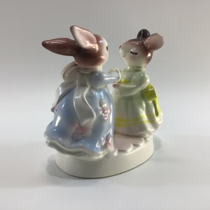 May include: A porcelain figurine of a rabbit and a mouse dressed in dresses, holding hands and facing each other. The rabbit is brown and white, wearing a blue dress with white trim and pink flowers. The mouse is brown and white, wearing a green dress with yellow trim and white flowers. The figurine is on a white base.
