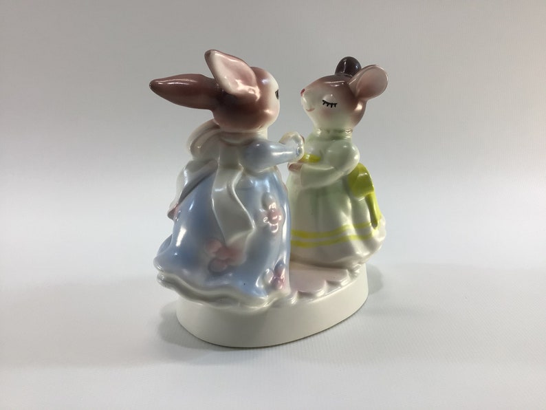 May include: A porcelain figurine of a rabbit and a mouse dressed in dresses, holding hands and facing each other. The rabbit is brown and white, wearing a blue dress with white trim and pink flowers. The mouse is brown and white, wearing a green dress with yellow trim and white flowers. The figurine is on a white base.