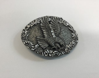 May include: A silver belt buckle with an eagle design. The eagle is in flight and has its wings spread wide. The buckle is surrounded by a floral design.