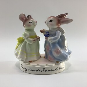 May include: A porcelain figurine of a mouse and a rabbit dressed in dresses, holding flowers. The base of the figurine reads "The day I made President's Club".