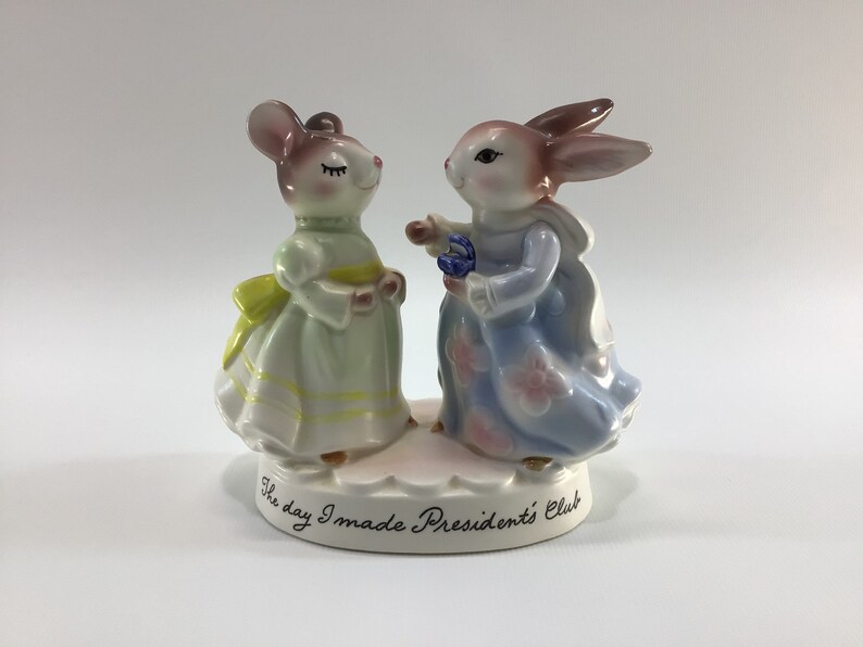 May include: A porcelain figurine of a mouse and a rabbit dressed in dresses, holding flowers. The base of the figurine reads "The day I made President's Club".