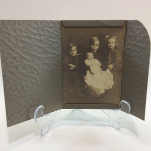 May include: A vintage photograph of four children, three standing and one sitting in a chair. The children are dressed in clothing from the early 1900s. The photo is in a brown cardboard frame.