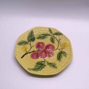 May include: A yellow ceramic plate with a woven texture and an octagonal shape. The plate is decorated with a branch of red and pink fruit, green leaves, and small yellow flowers.